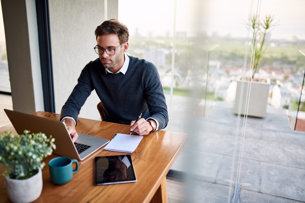 Homem dentro de empresa, sentado à uma mesa de escritório ou de reunião, usando notebook e bloco de anotações, representando a emissão de manifesto eletrônico MDFe