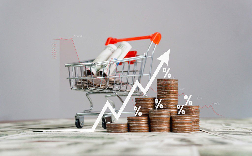 Miniatura de carrinho de compras com notas de dinheiro dentro, ao lado do carrinho pilhas de moedas de diferentes tamanhos, cada uma com um símbolo digital de porcentagem sobre a pilha e na imagem uma seta digital crescente para cima, passando por cima das moedas, simbolizando impostos sobre vendas