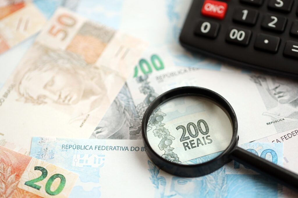 Notas de reais brasileiros e calculadora com lupa. Grande quantidade de notas de real brasileiro e calculadora com lupa sobre uma mesa. Conceito de tributação e contagem de dinheiro no Brasil.